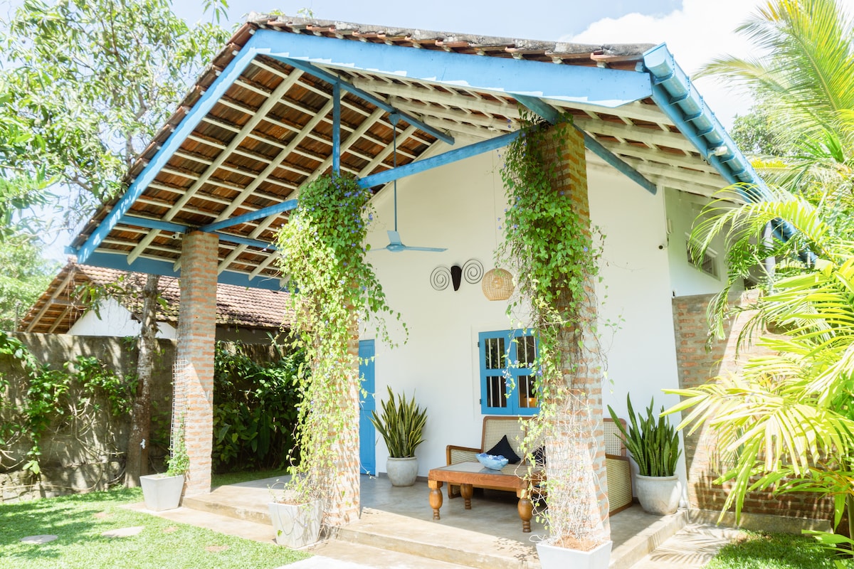 The entrance of the villa is framed by tall columns with climbing greenery, leading to a welcoming outdoor seating area. A wooden bench is positioned under a gently sloped roof, providing a shaded spot amidst gentle palm foliage and tropical plants.