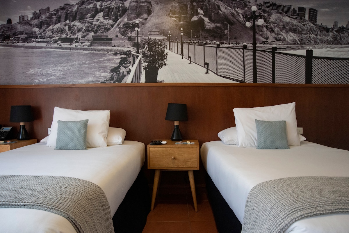 A serene guest room features two spacious beds with crisp white bedding and light grey throws. Each bed is flanked by wooden bedside tables topped with stylish lamps. A large black-and-white mural of a coastal scene adorns the wall, enhancing the room's calming ambiance.