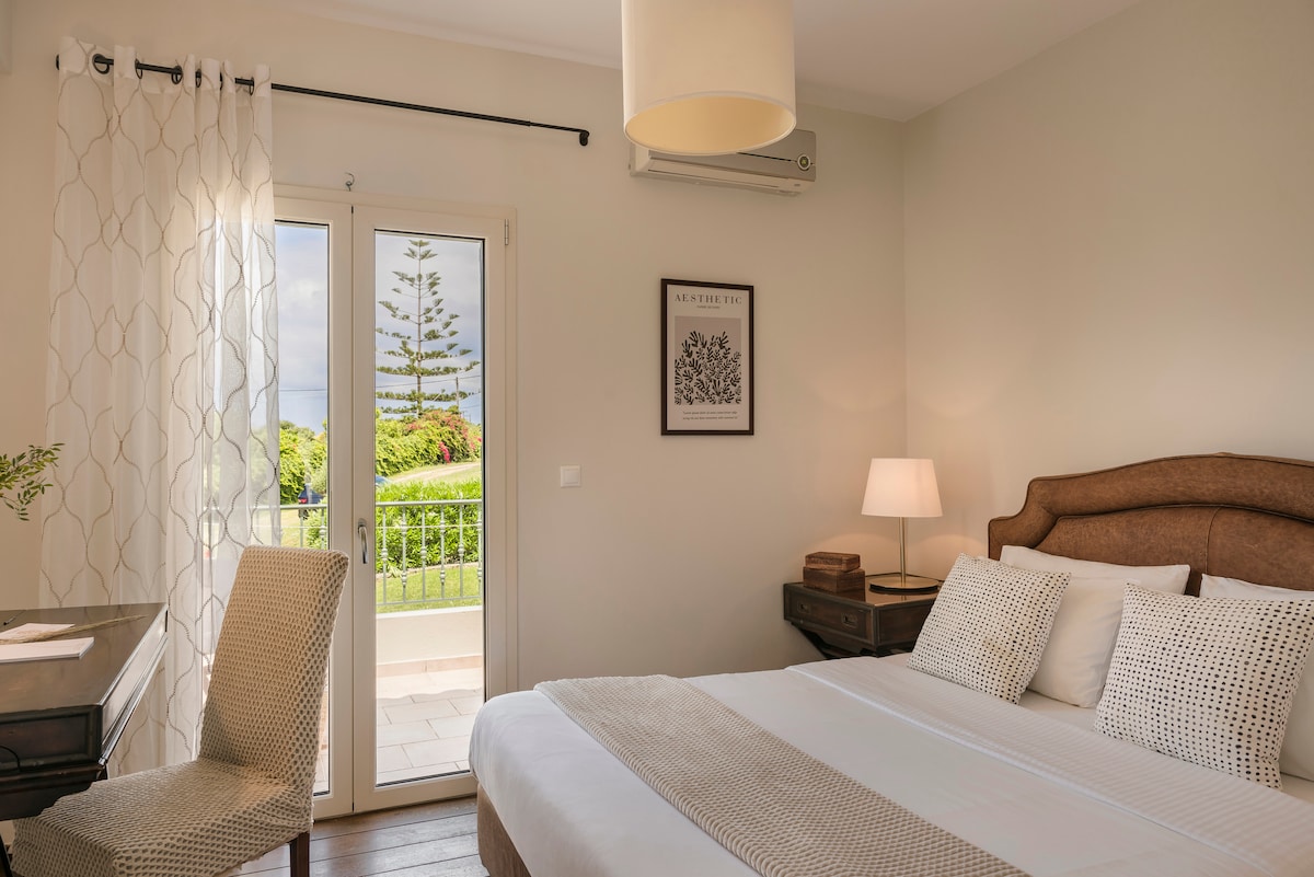 A serene bedroom features a spacious bed topped with neutral-toned linens, complemented by a wooden headboard. A desk with a chair is positioned near the window, which opens to a private balcony showcasing lush greenery and ocean views.