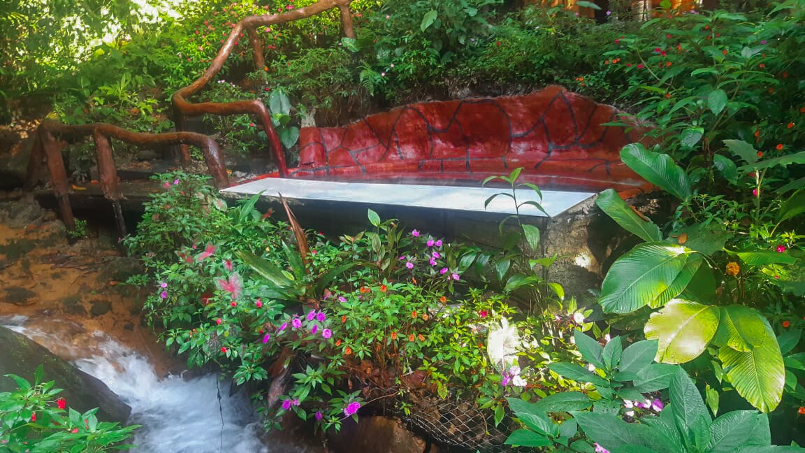 A tranquil seating area is nestled among lush greenery, featuring a stone bench adorned with vibrant flowers. A gentle stream flows nearby, its sound enhancing the peaceful ambiance of this natural retreat.