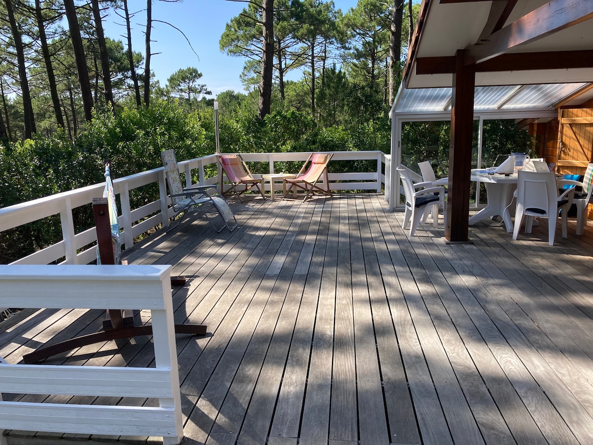 A spacious wooden terrace is surrounded by greenery, featuring two lounge chairs and a dining area with white furniture. The area is partially covered, and sunlight can be seen filtering through the trees, creating a calm and inviting outdoor space.