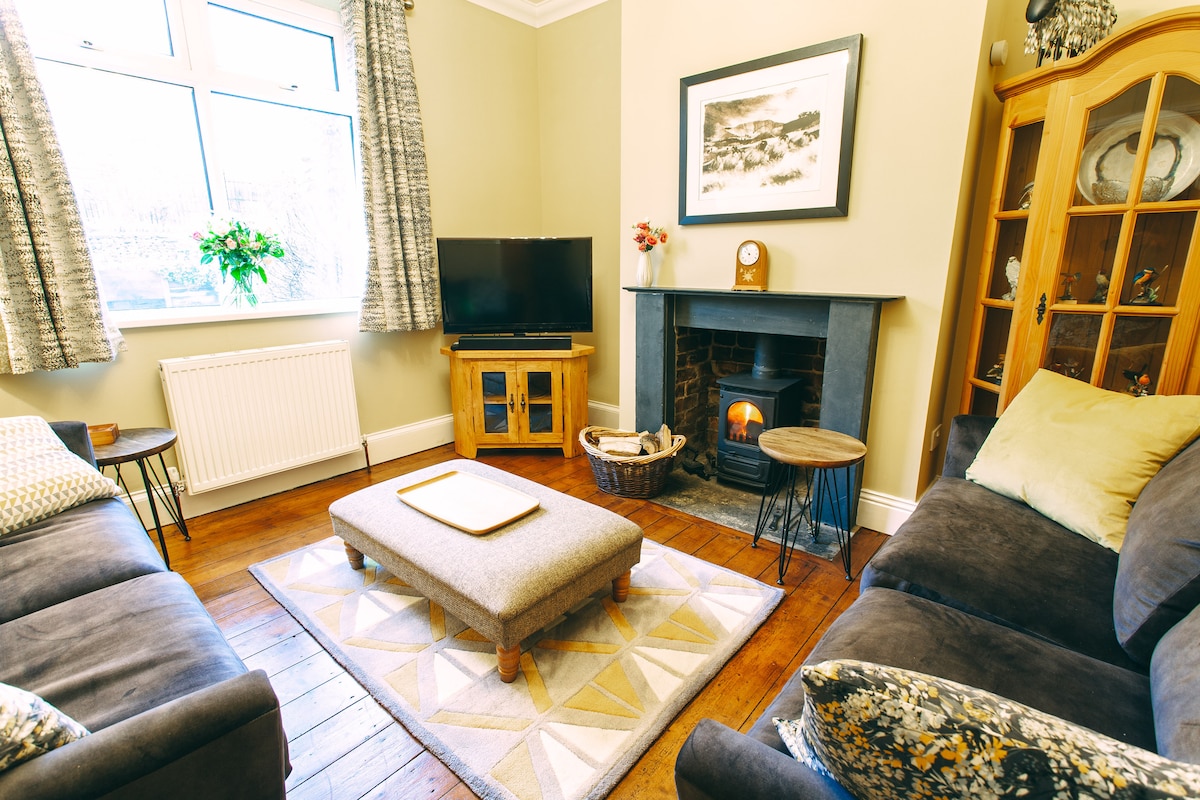 A comfortably arranged living room features dark sofas and a central ottoman on a patterned rug. A television is positioned opposite a wood-burning stove, accompanied by wooden side tables. Natural light enters through a large window adorned with patterned curtains.