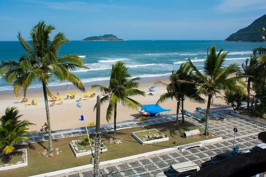 The image captures a beachfront view featuring a sandy beach bordered by palm trees. Beach chairs and umbrellas are arranged along the shore, while gentle waves can be seen lapping at the sand. The horizon presents a distant island against a clear blue sky.