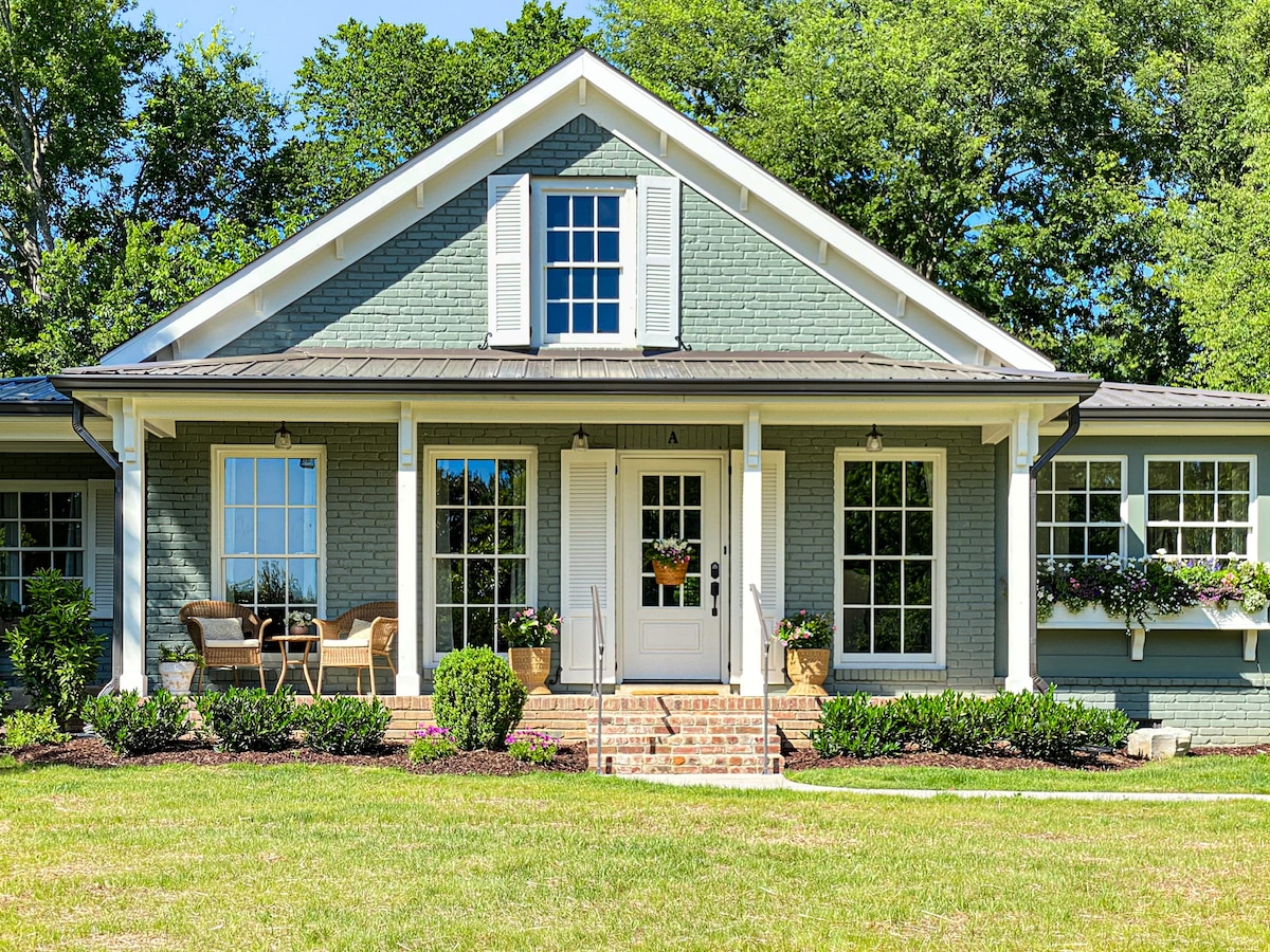 The exterior of a renovated cottage is showcased, featuring a light green brick facade and a symmetrical design. Large windows are framed with white shutters, while potted plants adorn the entrance. A welcoming porch includes two chairs and a decorative table, surrounded by landscaped greenery.