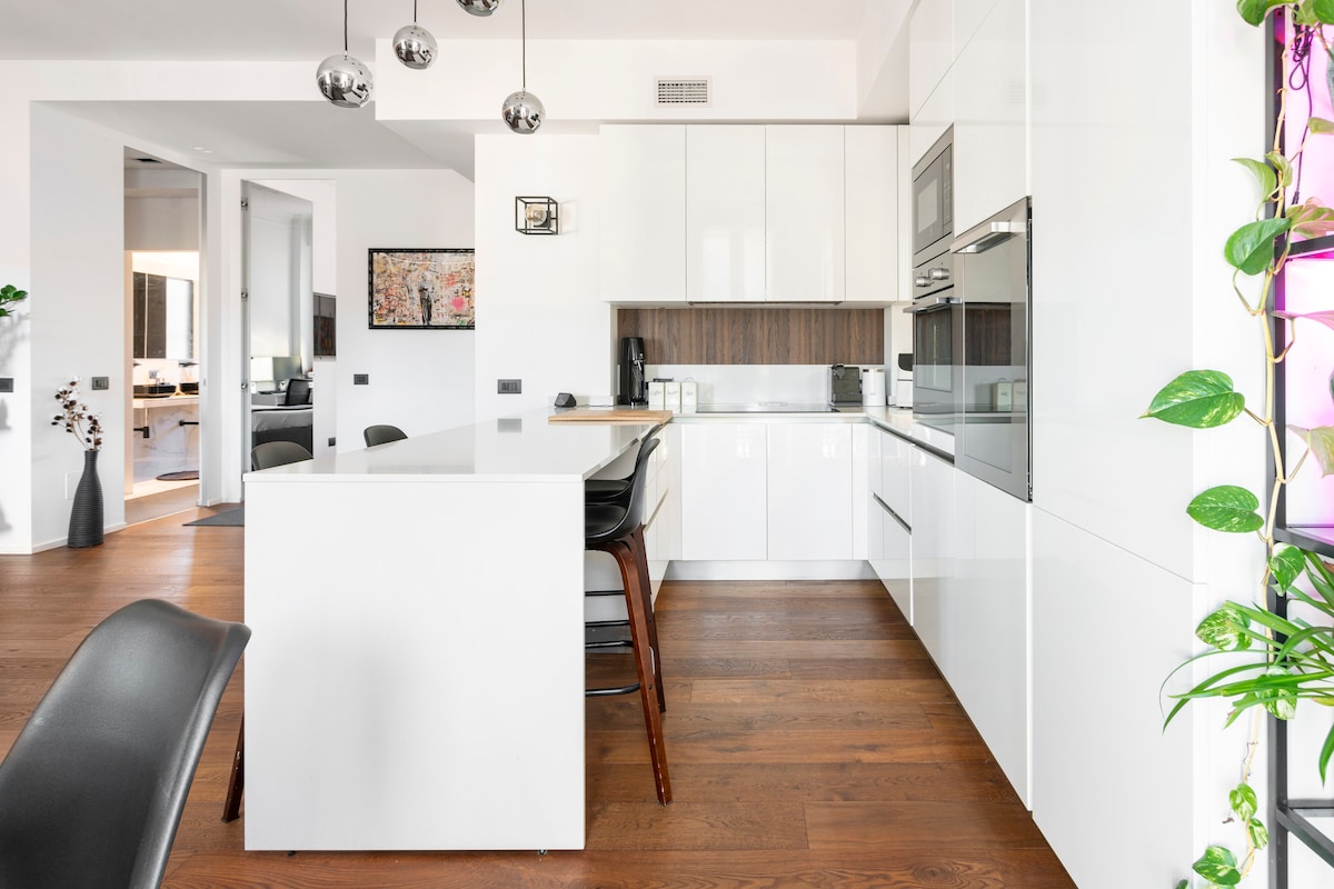 An airy kitchen features modern white cabinetry with wood accents and stainless steel appliances. A central island with seating is complemented by pendant lighting. The space is enhanced by natural light, hinting at the adjoining living areas visible through open doorways.