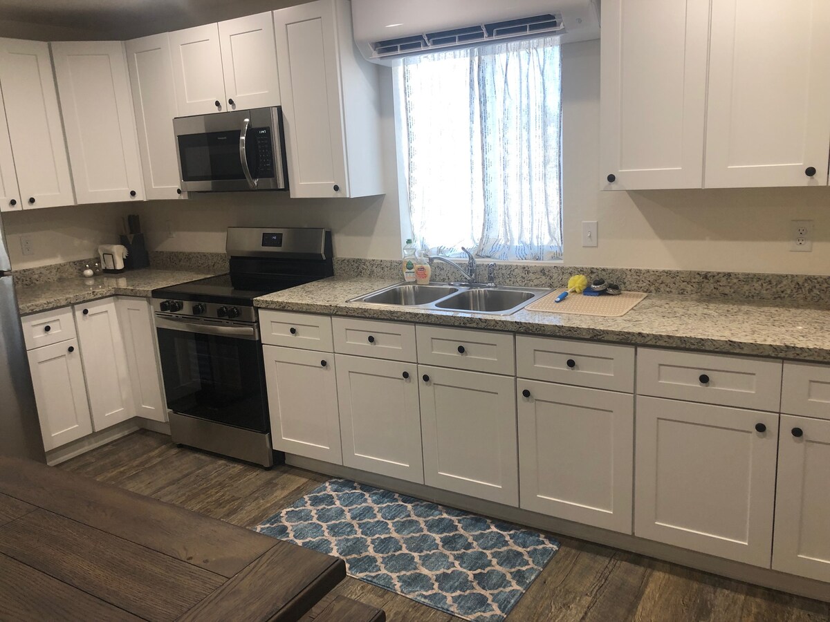 A well-equipped kitchen features white cabinetry and gray granite countertops. Stainless steel appliances, including an oven and microwave, are present. A double sink is positioned under a window framed with light curtains. A blue patterned rug adds a pop of color to the wooden floor.
