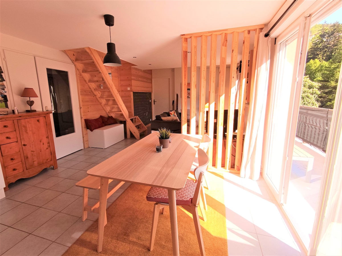 A bright dining area is illuminated by natural light, featuring a wooden table surrounded by four chairs. A decorative plant sits at the center. To the side, a wooden sideboard and framed windows allow views of the outdoors, complemented by a cozy seating area in the background.