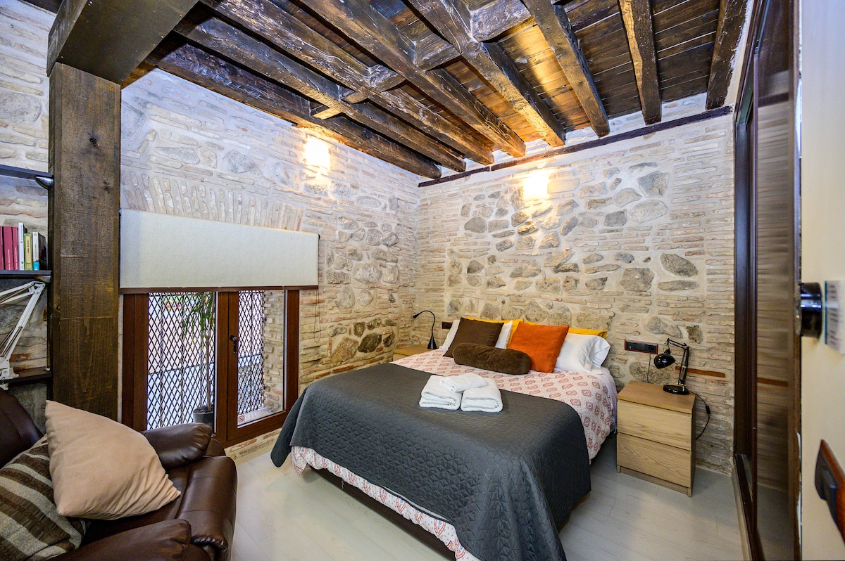 A cozy sleeping area features a spacious bed dressed in neutral and bright colors. Exposed wooden beams form the ceiling above, complementing the rustic stone walls. Warm light fixtures enhance the ambience, while a reading chair and bedside table provide additional comfort.