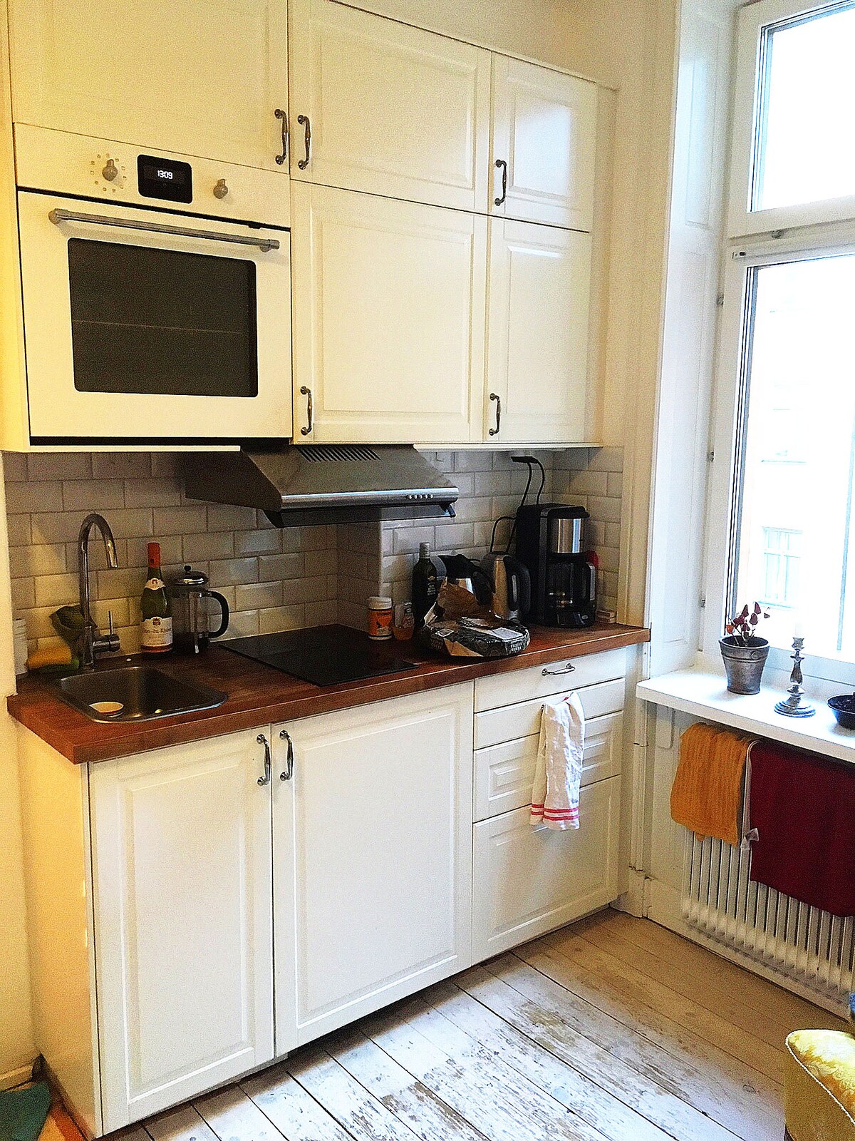 A compact kitchen area features white cabinetry and a wooden countertop. An oven and stovetop are built into the cabinetry, with a sink positioned beside them. A window allows natural light to enter, framed by curtains, while kitchen essentials and utensils are neatly organized.