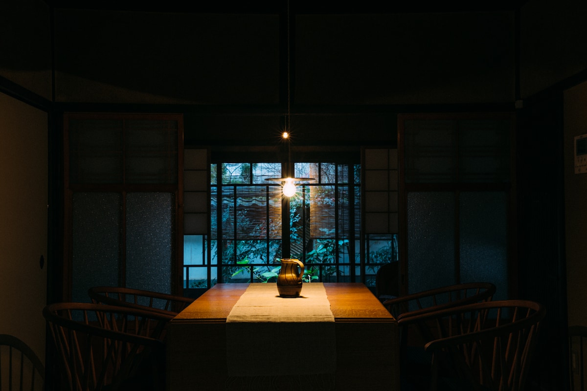 A dining area is illuminated by a pendant light, casting a warm glow over a simple wooden table adorned with a ceramic vase. The backdrop features a view of the garden through large sliding doors, framed by traditional shoji screens.