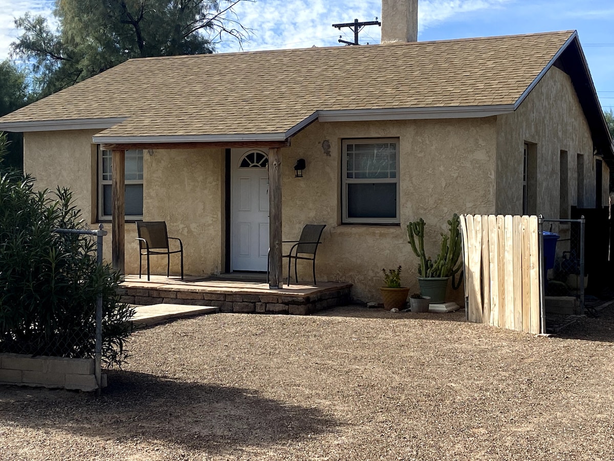 The exterior of the mud adobe home features a welcoming front porch with two chairs, framed by natural landscaping. The home's creamy facade contrasts with the brown gravel driveway, leading to a fenced area. A small potted plant is visible near the entrance.