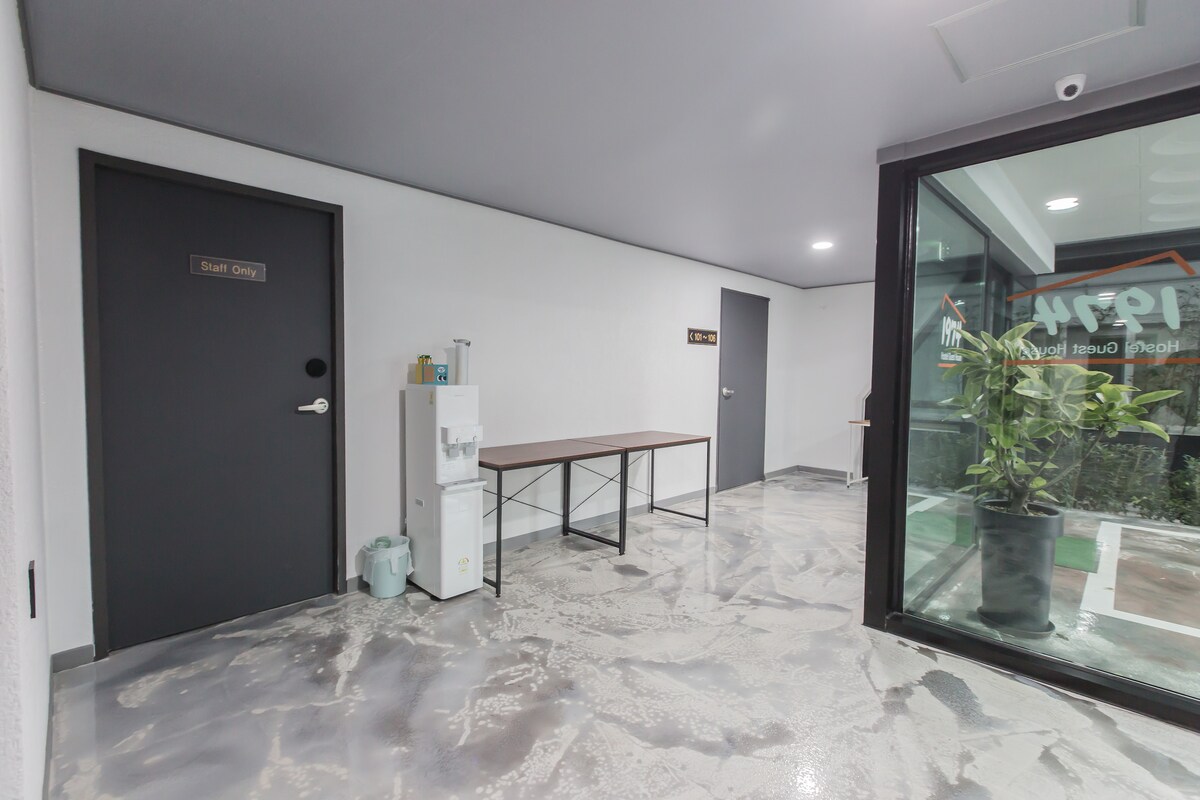 A clean and modern corridor is shown, featuring two closed doors labeled for staff and guests. A small table is positioned against the wall, complemented by a water dispenser nearby. Natural light streams in from a glass window, highlighting the polished concrete floor.