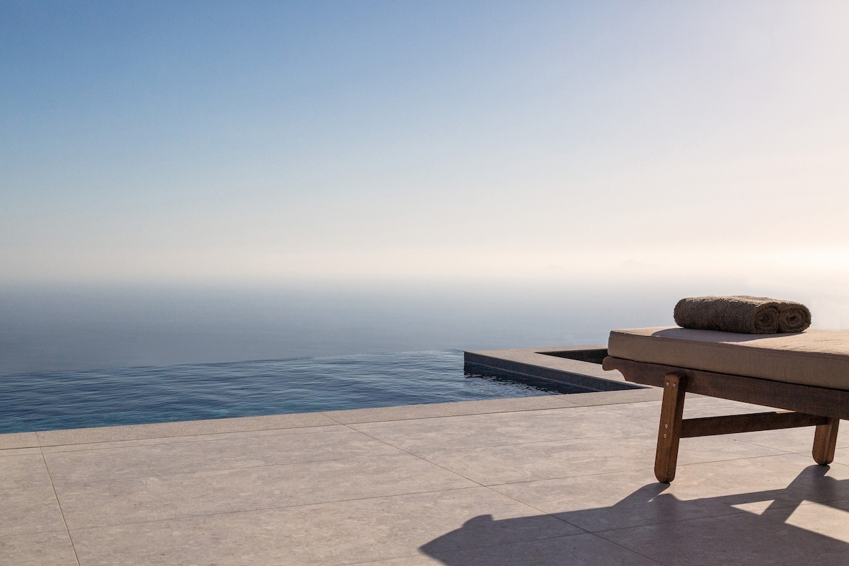 An outdoor terrace features a sun lounger positioned next to an infinity pool, extending towards the sea. Soft towels are rolled and placed on the lounger, while the horizon showcases a gentle blend of ocean blues under a clear sky.
