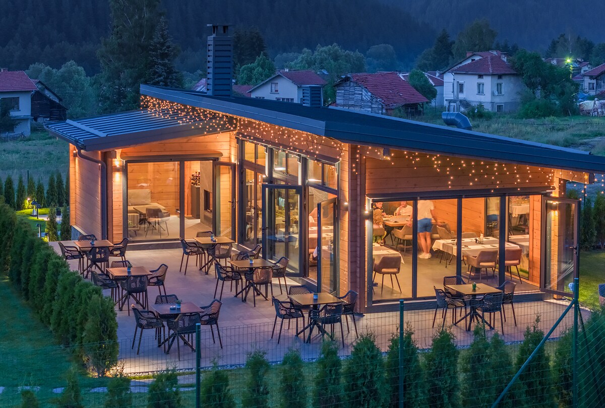 A modern chalet is featured, surrounded by greenery and illuminated by soft outdoor lighting. Tables and chairs are arranged on the patio, offering ample seating space. Large glass doors provide a view of the interior, where warm lighting enhances the inviting ambiance.