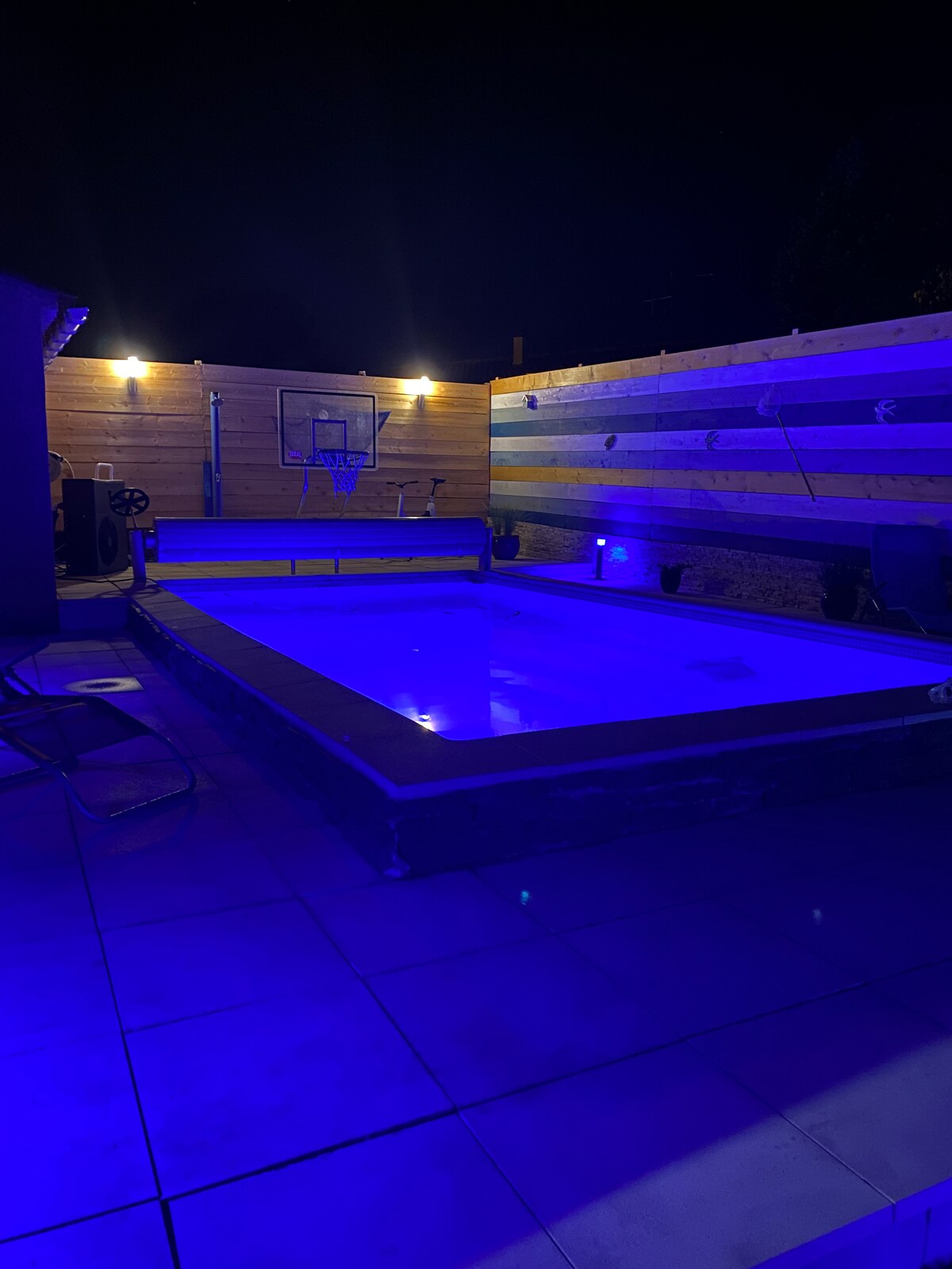 A rectangular swimming pool is illuminated in calming blue lights at night. Surrounding wooden panels provide privacy, while overhead lights enhance the ambiance. The pool area features tiled flooring, creating a cohesive outdoor space. A basketball hoop is positioned nearby, adding recreational options.