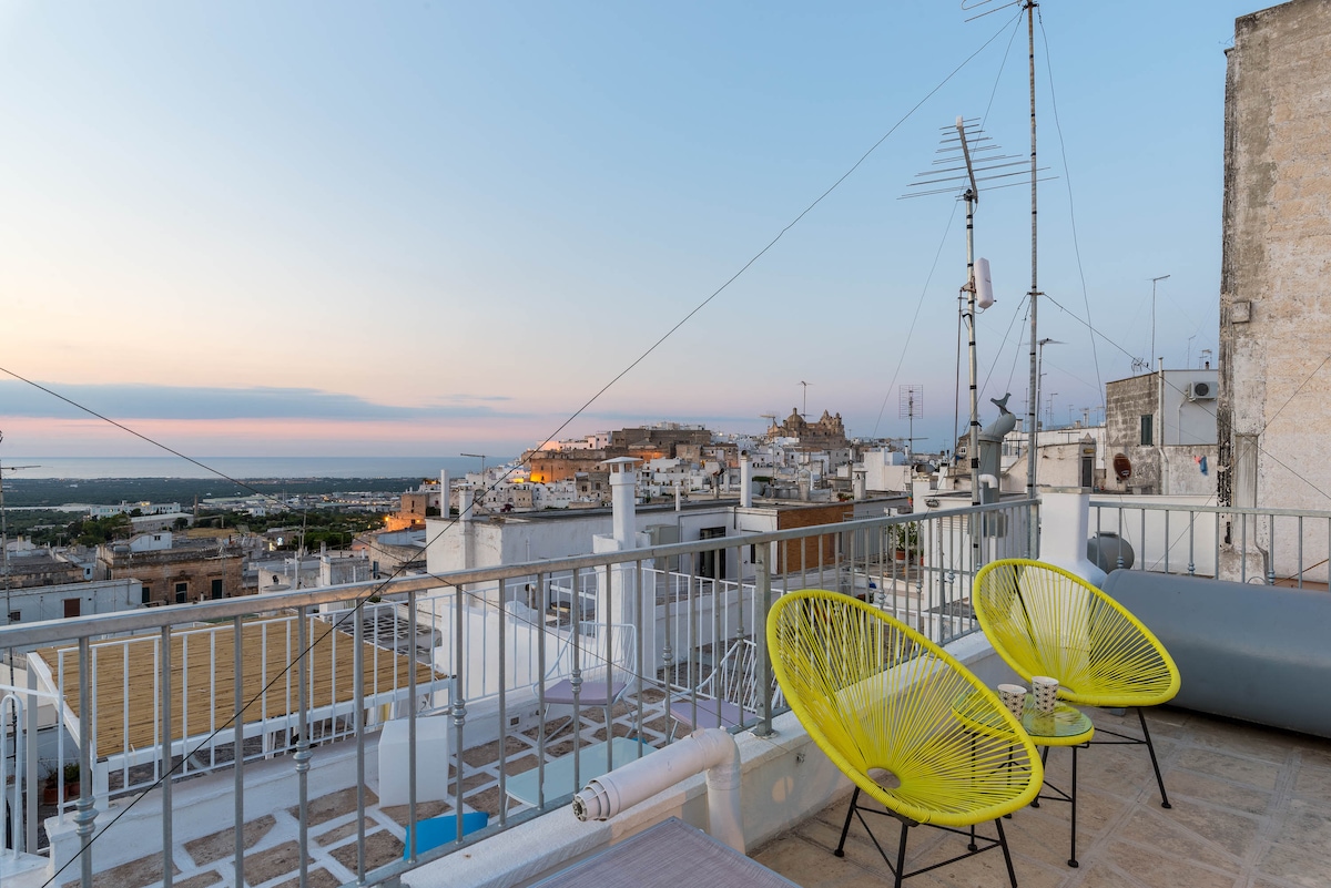 A private terrace offers a panoramic view of the surrounding whitewashed buildings and coastal landscape at sunset. Bright yellow chairs are positioned on the terrace alongside a small table, inviting guests to relax and enjoy the serene atmosphere.