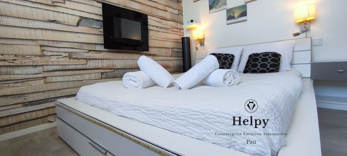 A spacious bed is dressed in white bedding, flanked by modern wall-mounted lamps. Towels are neatly arranged on the bed. A flat-screen TV is mounted on a wooden accent wall, contributing to a cozy setting.