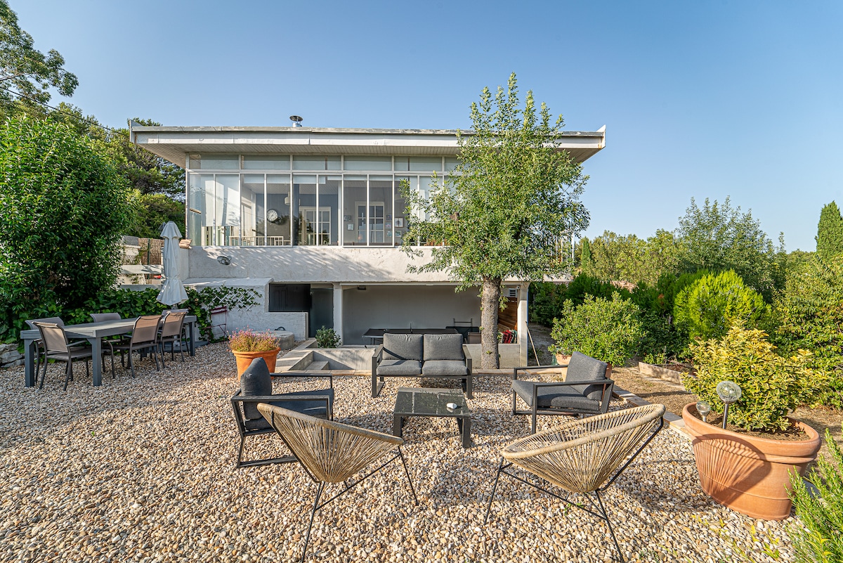 The exterior of the house reveals a spacious garden featuring several seating areas. A mix of lounge chairs and a dining table are set against a backdrop of lush greenery and decorative planters. Large windows of the house reflect the natural light, enhancing the inviting ambiance.