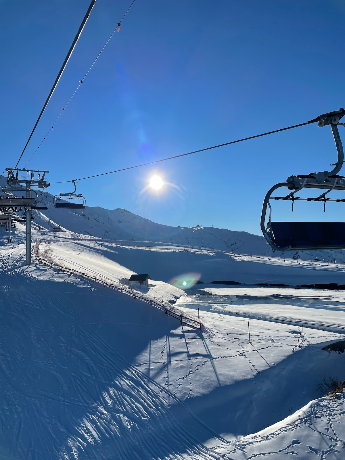 A view from a ski lift overlooking a snowy landscape illuminated by bright sunlight. The slopes are covered in fresh powder, with a distant cabin visible amidst the backdrop of mountains. The clear blue sky complements the winter scenery.