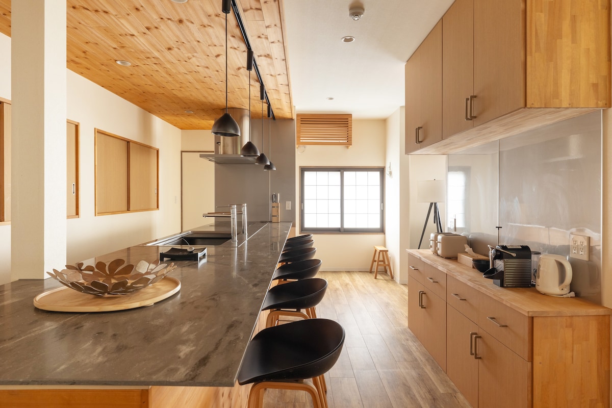 A spacious kitchen features a long, sleek counter with modern stools. The wood panel ceiling adds warmth to the neutral walls, while large windows allow natural light to fill the space. Appliances are neatly arranged along the counter, encouraging a communal cooking experience.