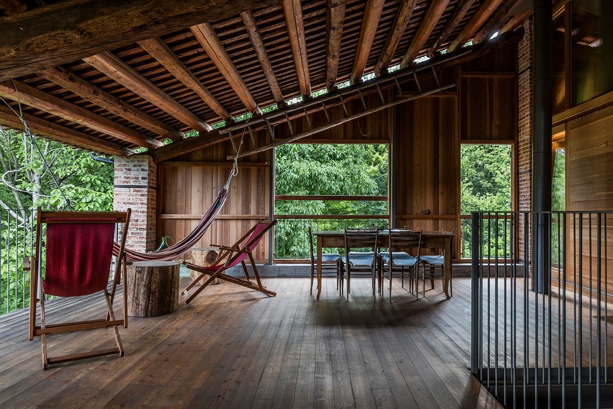 An open wooden space features high ceilings with exposed beams. Large windows allow views of the surrounding greenery. A hammock hangs between two supports, while a wooden dining table and chairs occupy one side, inviting relaxation and connection with nature.