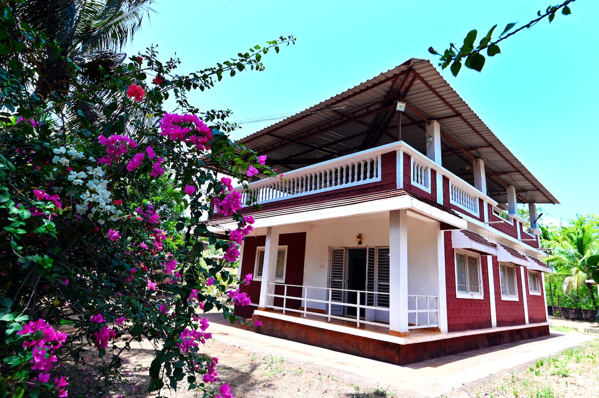 A two-story eco-friendly home is surrounded by vibrant flowering plants. The building features a spacious balcony with white railings, large windows, and a red exterior, harmonizing with the lush greenery of the environment.