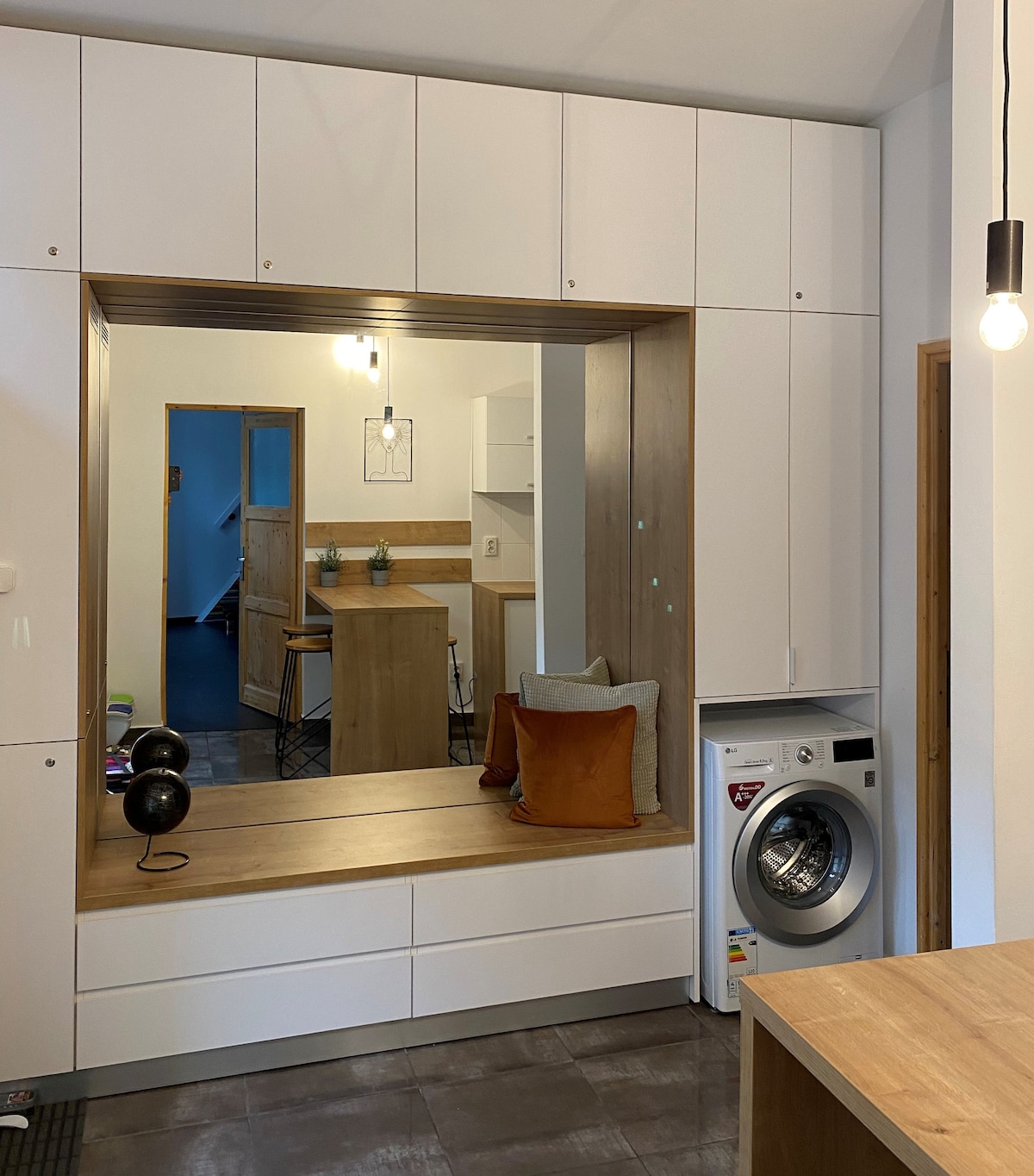 A modern kitchen area is highlighted by minimalist cabinetry and a central island with seating. A washing machine is tucked discreetly next to the cabinetry. Warm lighting hangs above, casting a gentle glow across the space, which features neutral tones and sleek surfaces.