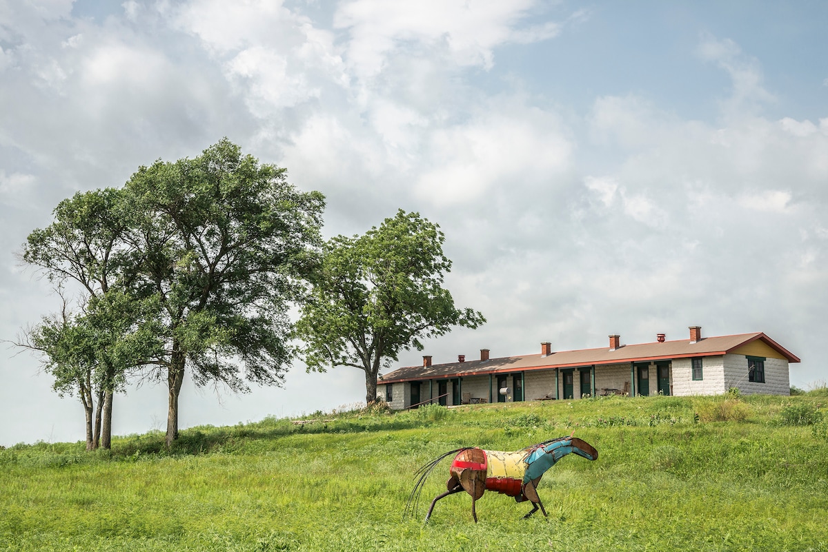 A historic lodging structure is set against a cloudy sky, nestled among green grassy fields. Two large trees flank the building, while a colorful horse sculpture adds artistic flair to the landscape, contributing to the serene rural atmosphere.
