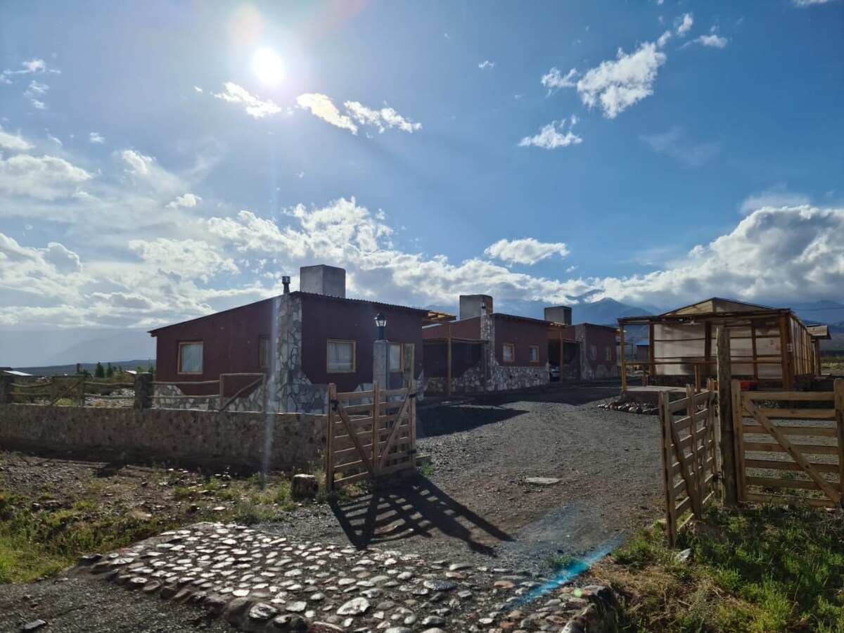 A rustic building made of stone and wood is depicted, set against a bright blue sky with scattered clouds. The sun shines down, illuminating the entrance and surrounding gravel pathway, leading through a welcoming gate towards the structure.