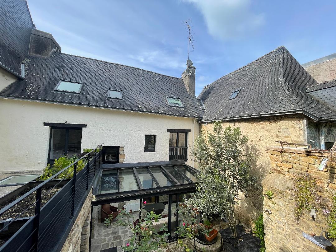 An outdoor view showcases the charming architecture of the stone building, complemented by a modern glass structure. Multiple skylights are visible along the roofline, and greenery is seen in the courtyard, enhancing the tranquil atmosphere of the space.