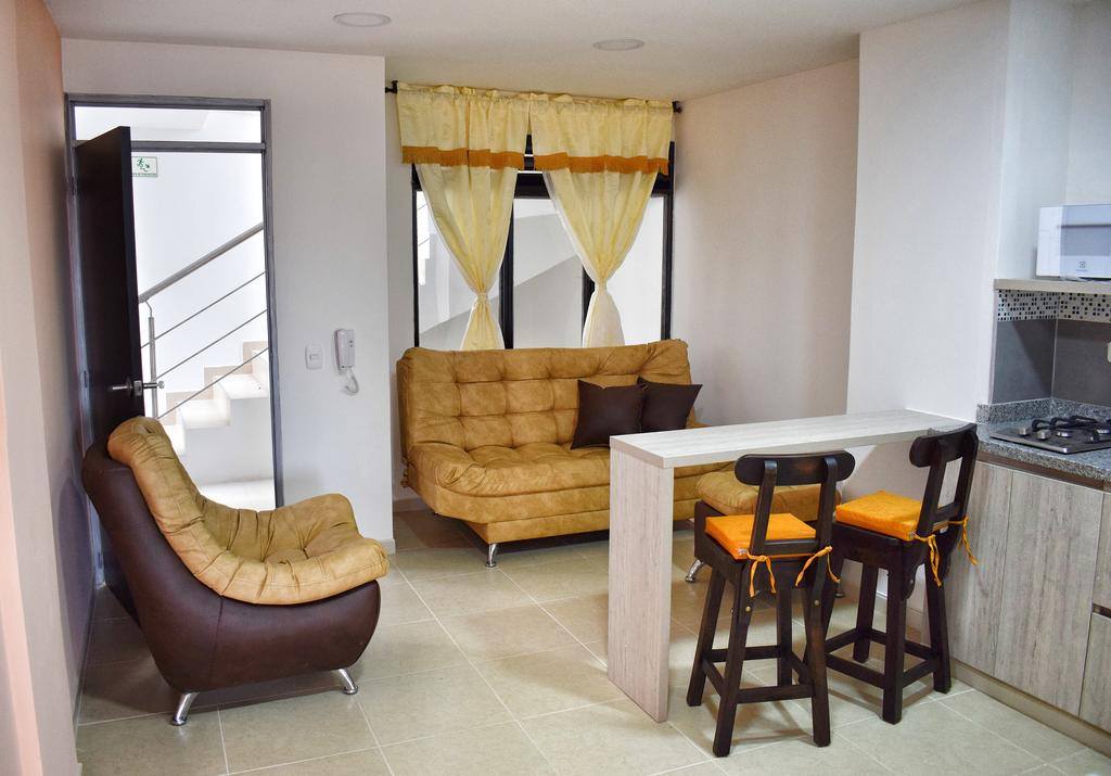 A cozy living area is featured with a soft tan sofa and a brown accent chair. A small dining table with two stools is adjacent to a kitchenette. Natural light enters through large windows dressed with sheer yellow curtains.