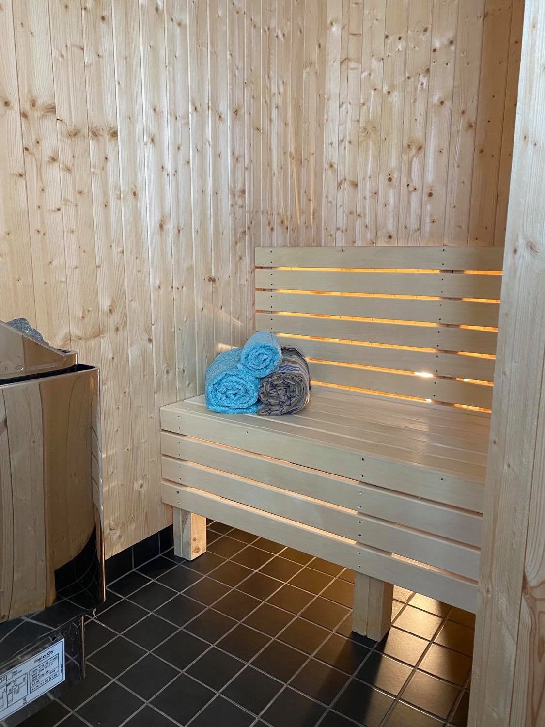 A wooden sauna area features a slatted wooden bench illuminated by soft light. Rolled towels in shades of blue and grey are neatly placed on the bench, providing an inviting touch to the calming space. The flooring is dark tiled, adding contrast to the natural wood.
