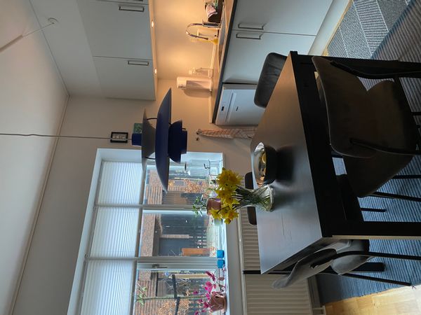 A well-lit kitchen area features a dark table surrounded by four upholstered chairs. Natural light enters through a large window adorned with white blinds. A pendant light hangs above the table, while a vase with yellow flowers and a bowl of fruit sits centrally on the table.