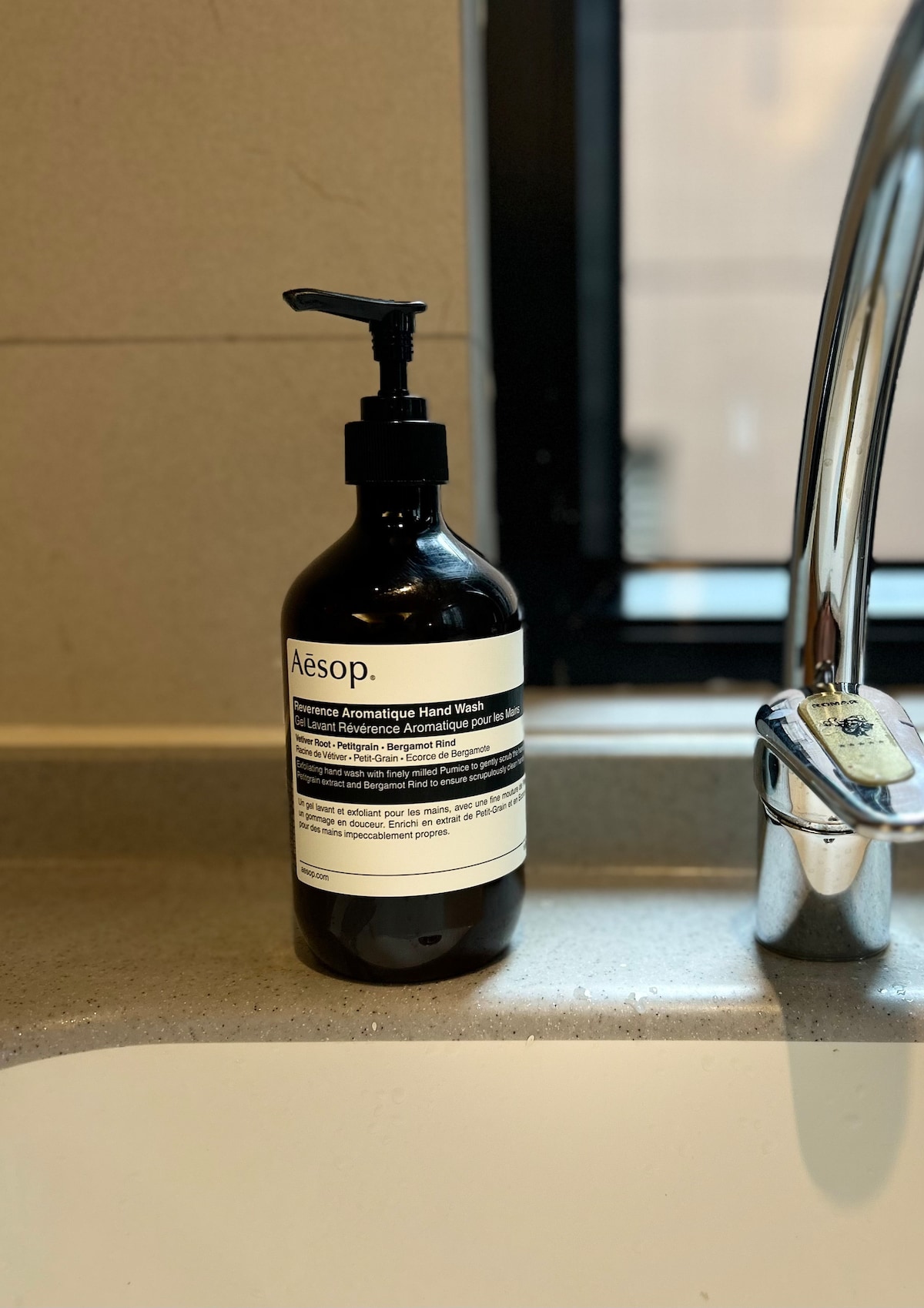 A bottle of Aesop hand wash is placed near a modern sink. The dark glass bottle features a pump dispenser and a label with product information. A sleek faucet reflects light, while the countertop is finished in light-colored stone.