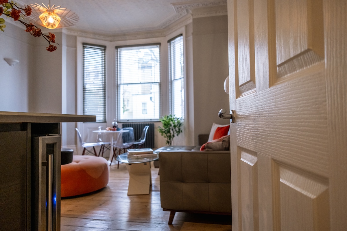 A view of the living area is presented, featuring large windows that fill the space with natural light. A comfortable sofa is positioned next to a cozy orange pouf. A small dining table with chairs is visible in the background, along with greenery from potted plants.