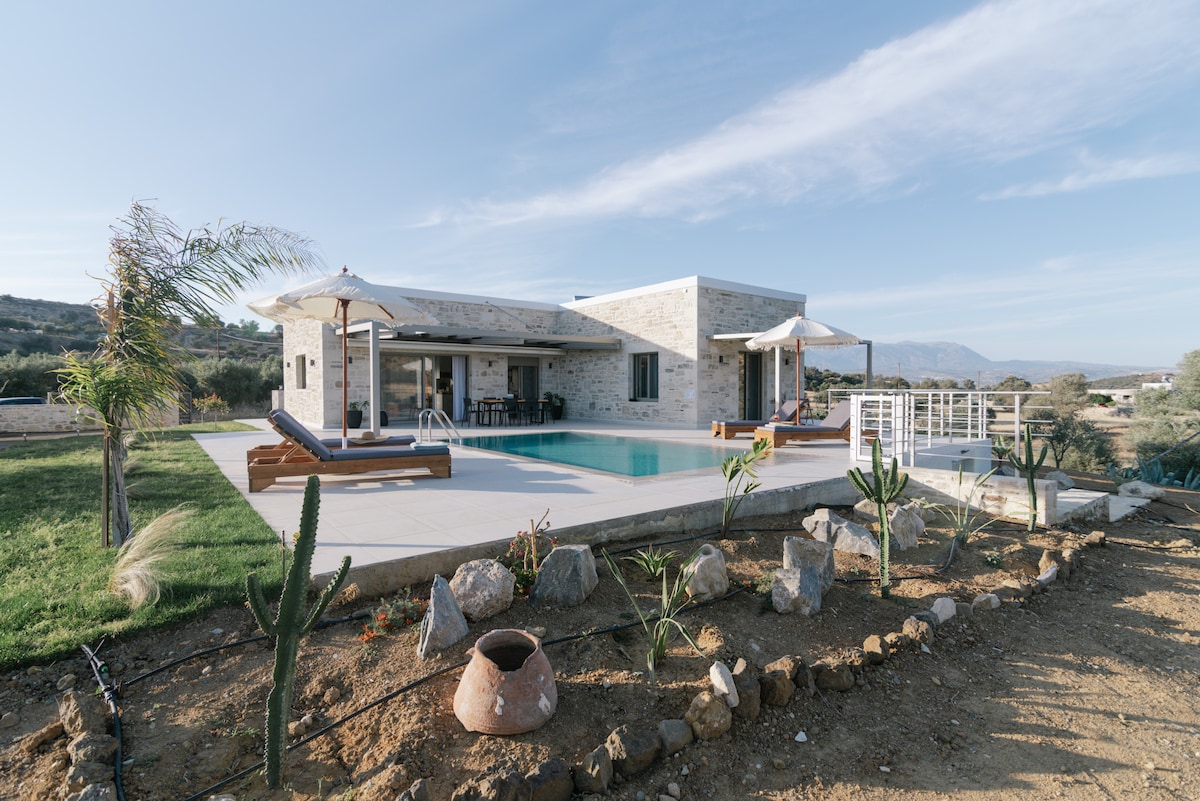 The modern villa features large sliding glass doors that open to a sunlit patio area with a swimming pool. Two umbrellas provide shade for outdoor seating beside the pool. Lush greenery and strategically placed rocks outline the yard, enhancing the serene environment.