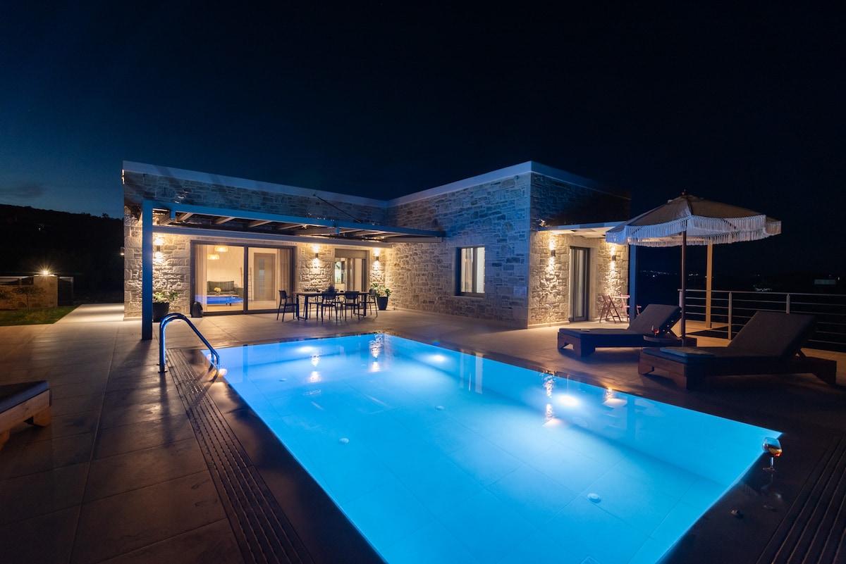 The villa exterior is illuminated at night, highlighting a modern design complemented by a sparkling blue pool. Comfortable lounge chairs sit beside the pool, while a patio area features a dining table under a covered terrace, creating a serene outdoor setting.