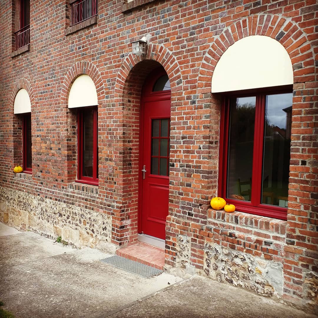 The entrance is showcased with a vibrant red door, framed by warm brick walls featuring three tall windows with elegant arches. Small pumpkins are placed on the ledge below the windows, adding a seasonal touch to the inviting facade.