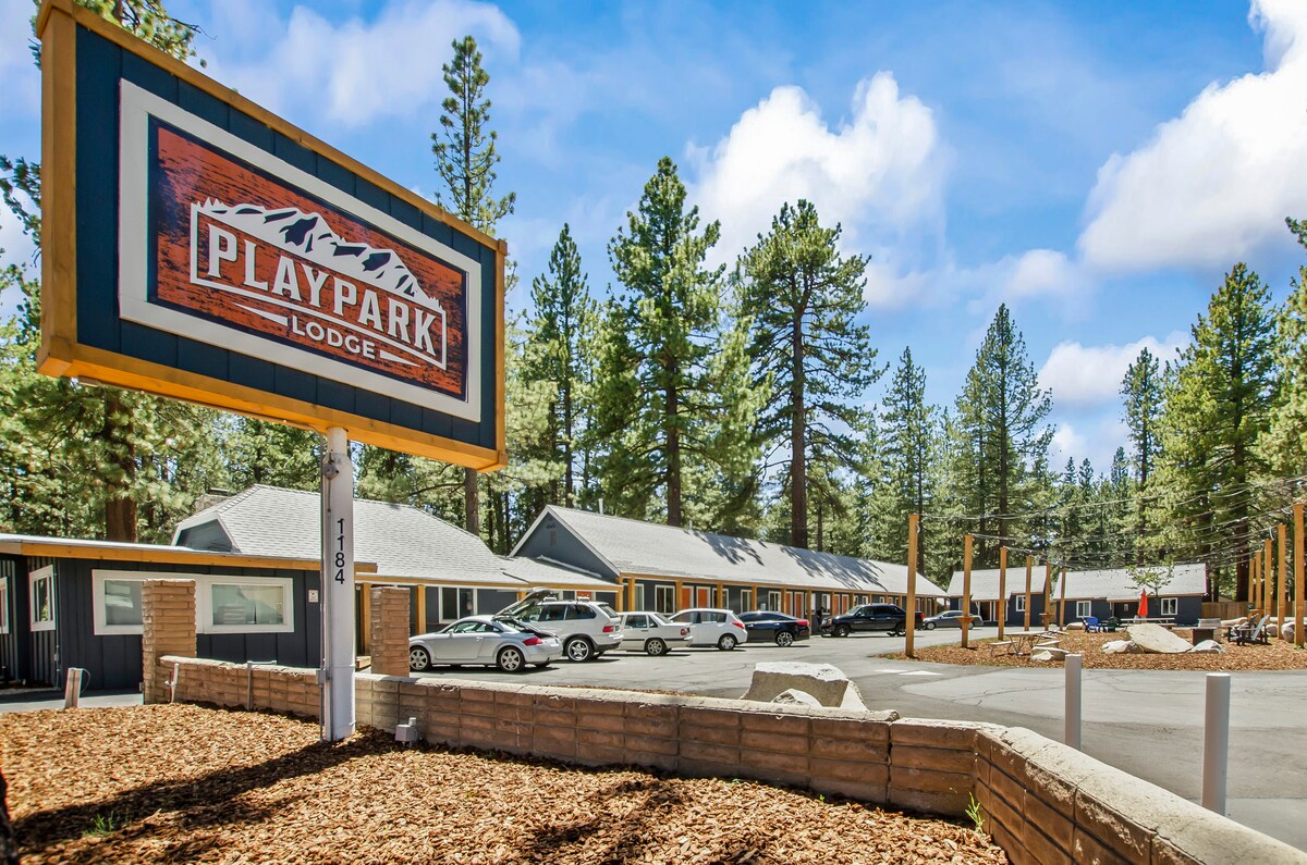 The exterior of the Playpark Lodge is displayed, featuring a large, welcoming sign. Surrounding trees provide natural shade, and well-maintained parking spaces are visible. The lodge structure includes multiple rooms with a modern facade, situated in a tranquil, forested area.