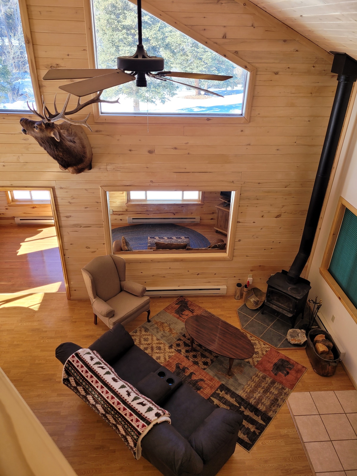 A spacious living area features a comfortable sofa adorned with a patterned blanket. A wooden coffee table is centered on a colorful area rug. Large windows bring in natural light, while a ceiling fan and a wood stove contribute to the cabin's cozy ambiance.