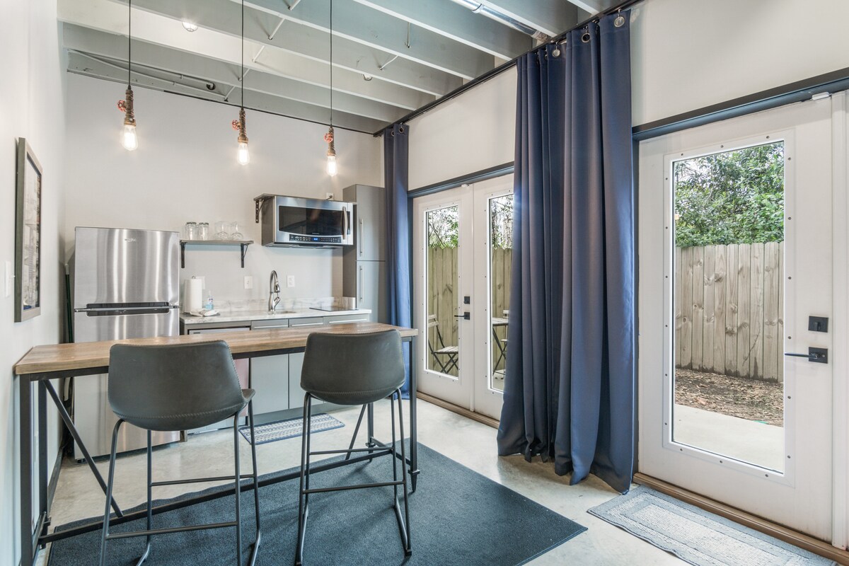 A modern kitchenette is presented with elegant cabinetry and stainless steel appliances. Two bar stools are positioned at a wooden island, while large glass doors provide access to the private outdoor space, framed by flowing dark curtains and minimalist decor.