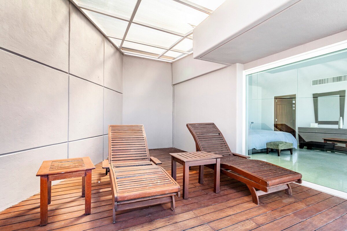 A private terrace features two wooden lounge chairs and a small table, all positioned on a wooden deck. Natural light fills the space, with glass doors leading to an interior room that contains a bed, seen in the background.