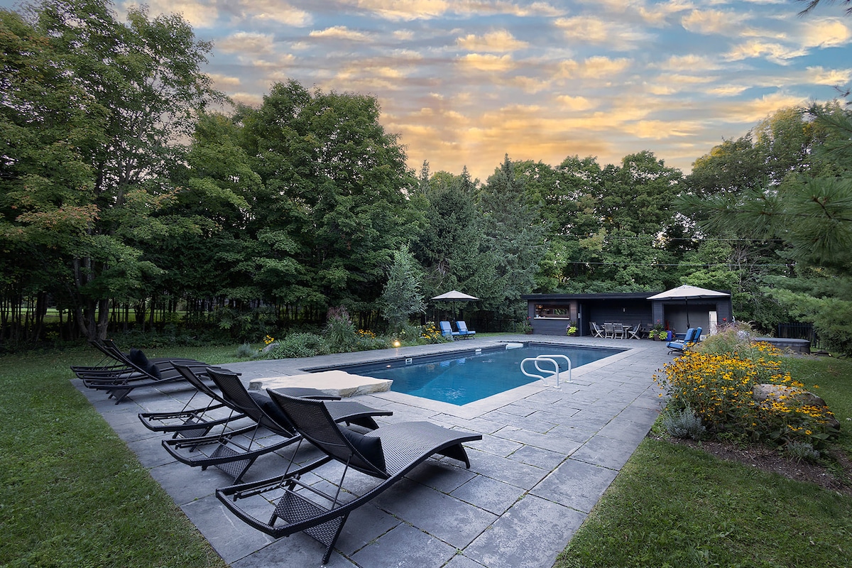 A spacious salt-water swimming pool is framed by lush greenery and vibrant flower beds. Lounge chairs are arranged around the pool area. Soft evening light casts a serene glow over the scene, complemented by the partly cloudy sky above.