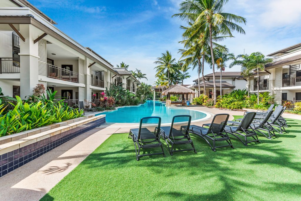 The image captures a serene pool area surrounded by tropical foliage. Several lounge chairs are positioned on lush green grass, offering a relaxing space. In the background, palm trees sway beside the resort buildings, and a thatched-roof gazebo can be seen near the water's edge.