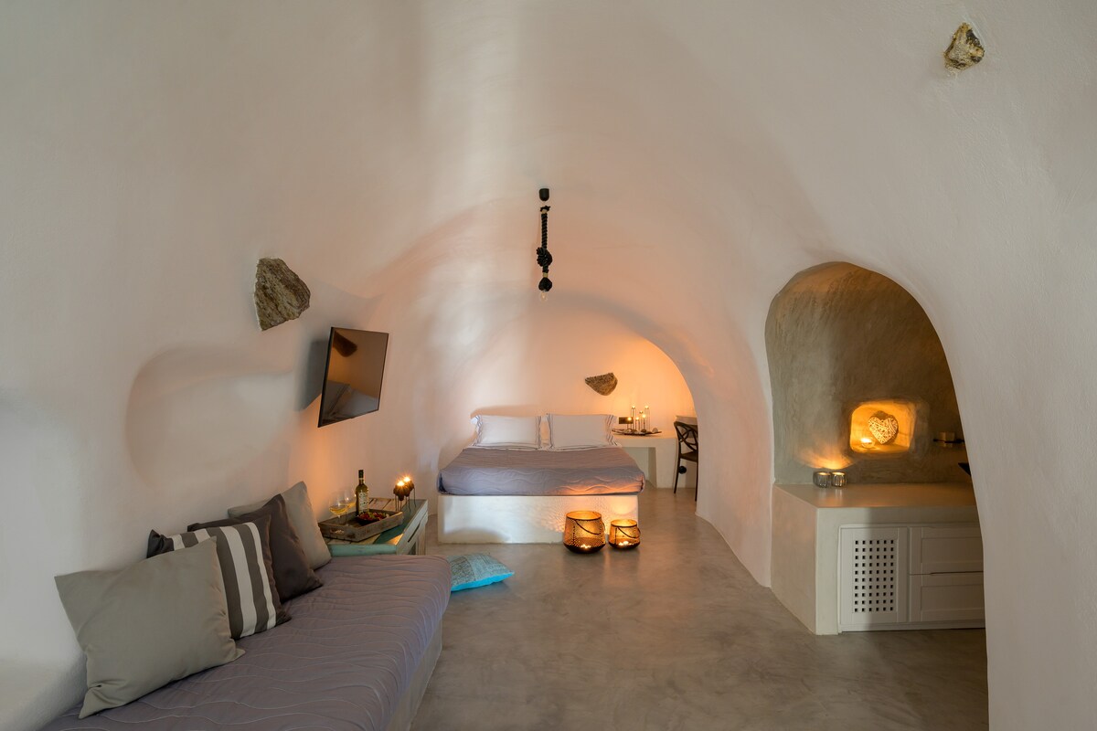 The interior of the cave villa features a spacious sleeping area with soft lighting and a comfortable bed. A cozy seating area includes a sofa, while a small kitchenette is visible in the background, with natural stone elements adding character to the space.