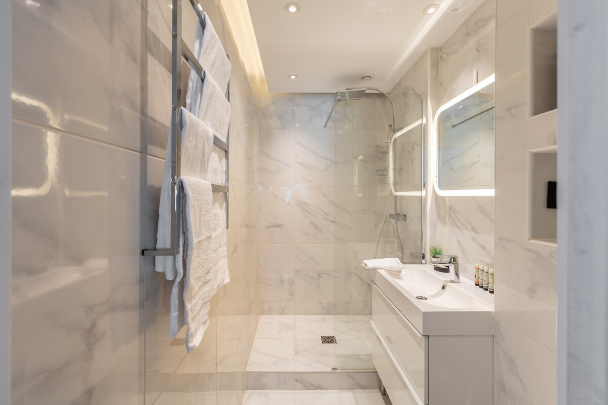 The bathroom features modern fixtures and marble finishes, with a spacious walk-in shower equipped with a rainfall showerhead. A sleek vanity with ample storage sits across from a wall-mounted mirror, complemented by soft lighting. Fresh towels hang neatly on a heated towel rail.