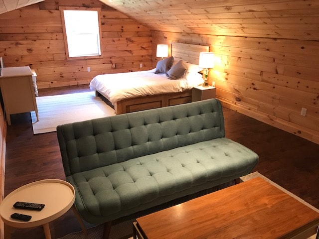 A spacious loft area features warm wooden walls and a large bed with neutral bedding. A light blue couch is positioned in the foreground, accompanied by a small round table and a wooden coffee table. Soft lighting is provided by bedside lamps.