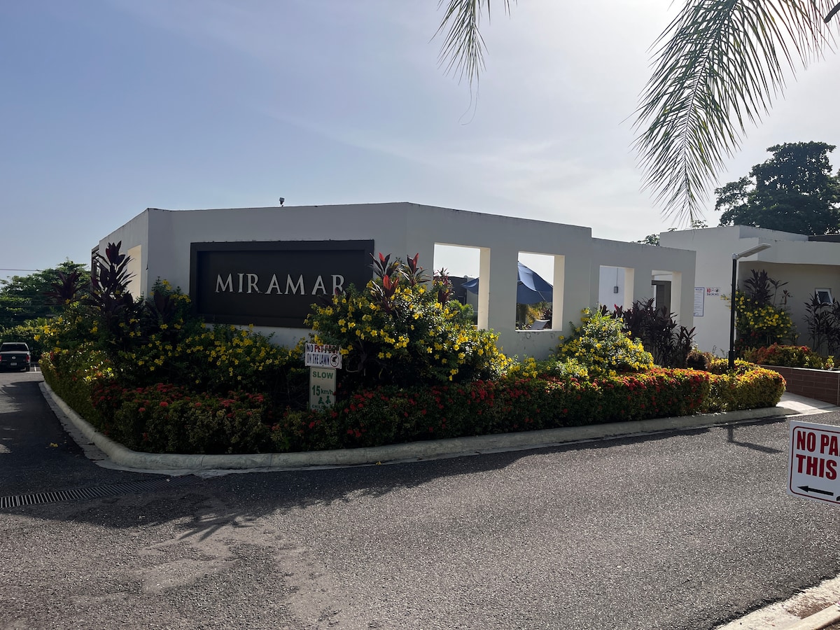 The entrance to the Miramar condominium is framed by vibrant flowers and lush greenery. A sign displaying the name 'MIRAMAR' is prominently featured, while a clear blue sky can be seen above. The pathway is bordered by neatly arranged shrubs.