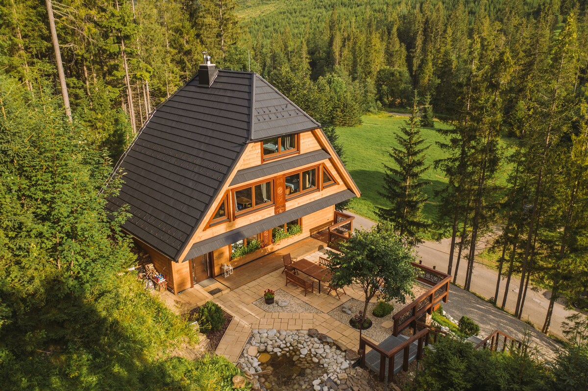 An aerial view of a chalet surrounded by lush greenery, featuring a dark sloped roof and large front windows. The stone pathway leads to a terrace with outdoor seating and a small garden. Dense forest borders the property, providing a serene backdrop.