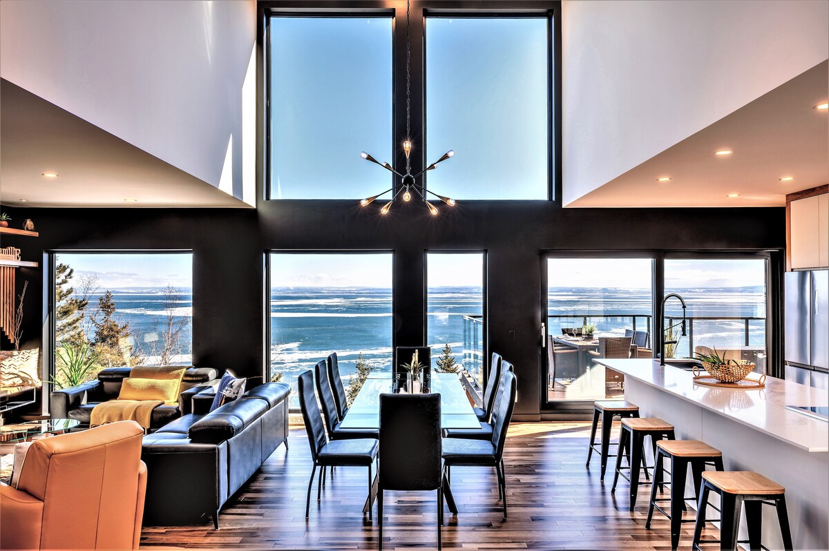 A spacious open-plan living area features large windows offering unobstructed views of the St. Lawrence River. A modern dining set is positioned beside the windows, while a cozy seating area with a sleek couch invites relaxation. Natural light highlights the elegant interior design.